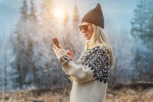 Outdoor close up portrait of young beautiful woman with long hair wearing hat, sweater posing with her phone in nature view. Christmas, winter holidays concept. Snowfall. Snow trees on background.