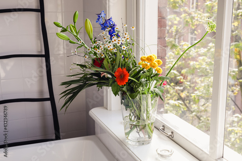 A bouquet of fresh spring wildflowers in a vase standing on a window sill in a big bright bathroom on a sunny day. Cozy home interior decor.