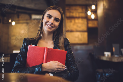 Half length portrait of cheerful hipster girl with literature book in hands smiling at camera, funny Caucaisasn female student holding education textbook and posing during leisure pastime indoors