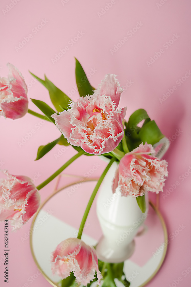 A bouquet of fresh parrot tulips with green leaves in a white ceramic jug standing on a round mirror against pink paper backdrop. Flower geometric decorative composition.