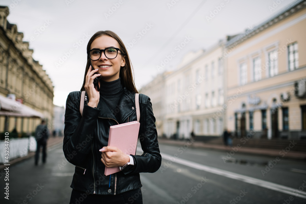 Fototapeta premium Millennial female student with education textbook using roaming internet connection for phone talking, happy tourist in optical eyewear making international conversation via smartphone application