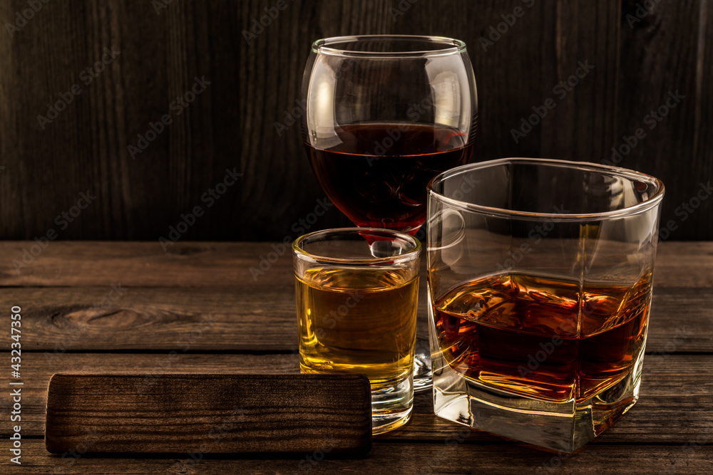 Three glasses with brandy, tequila and red wine with the empty wooden plank on an old wooden table. Angle view focus on the wooden plank Three glasses with brandy, tequila and red wine with the empty wooden plank on an old wooden table. Angle view focus on the wooden plank