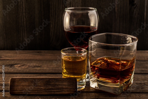 Three glasses with brandy, tequila and red wine with the empty wooden plank on an old wooden table. Angle view focus on the wooden plank