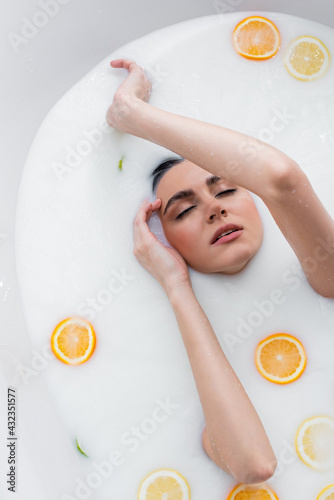 top view of woman with closed eyes bathing in milk with sliced citruses.