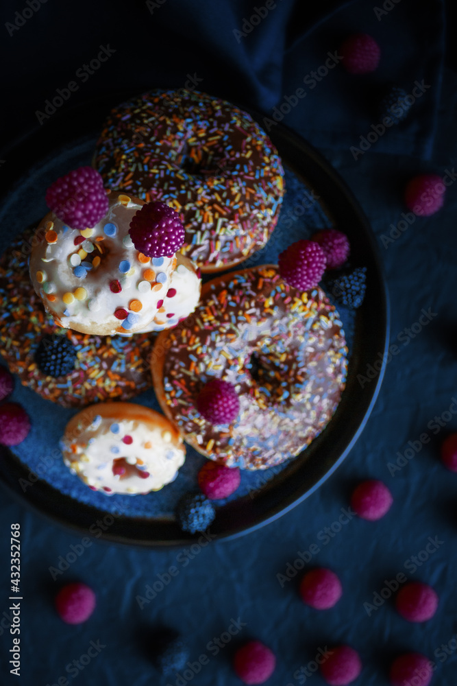 White and brown donuts decorated sprinkles on a blue plate, pink and black gelatin raspberry, levitation,  low-key lighting, flat lay, low-key lighting