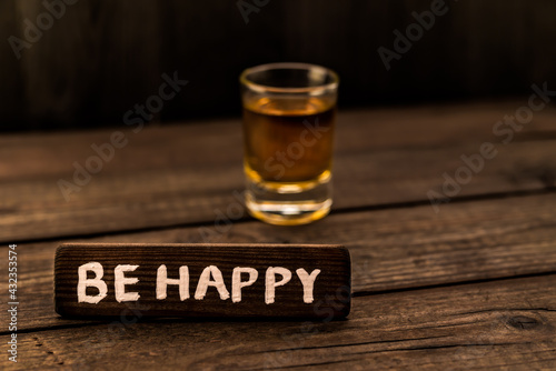 Glass of tequila with the wooden plank on it is an inscription "BE HAPPY" on an old wooden table. Angle view, focus on the inscription