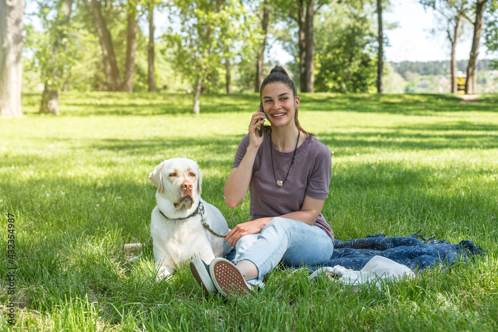 Young beautiful woman talking on her smartphone while is playing and enjoying with her pet dog the golden Labrador Retriever in the park on a sunny and beautiful day