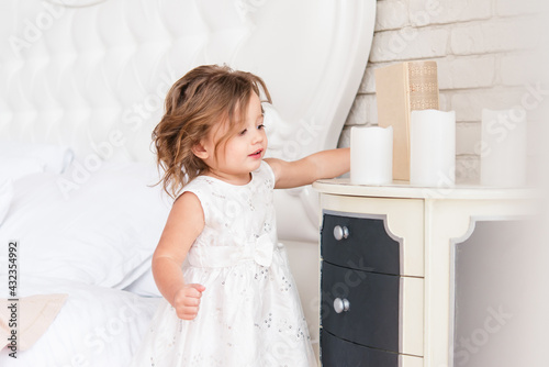 Cute little girl stands near the dresser. She is wearing a simple white dress. Bright room. European race.