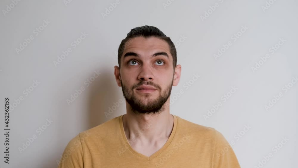 Human in yellow Tshirt on white studio background depicts emotions. Handsome Caucasian man with beard and ponytail on his head looks forward carefully, then looks up in surprise and blinks. Human in yellow Tshirt on white studio background depicts emotions. Handsome Caucasian man with beard and ponytail on his head looks forward carefully, then looks up in surprise and blinks.