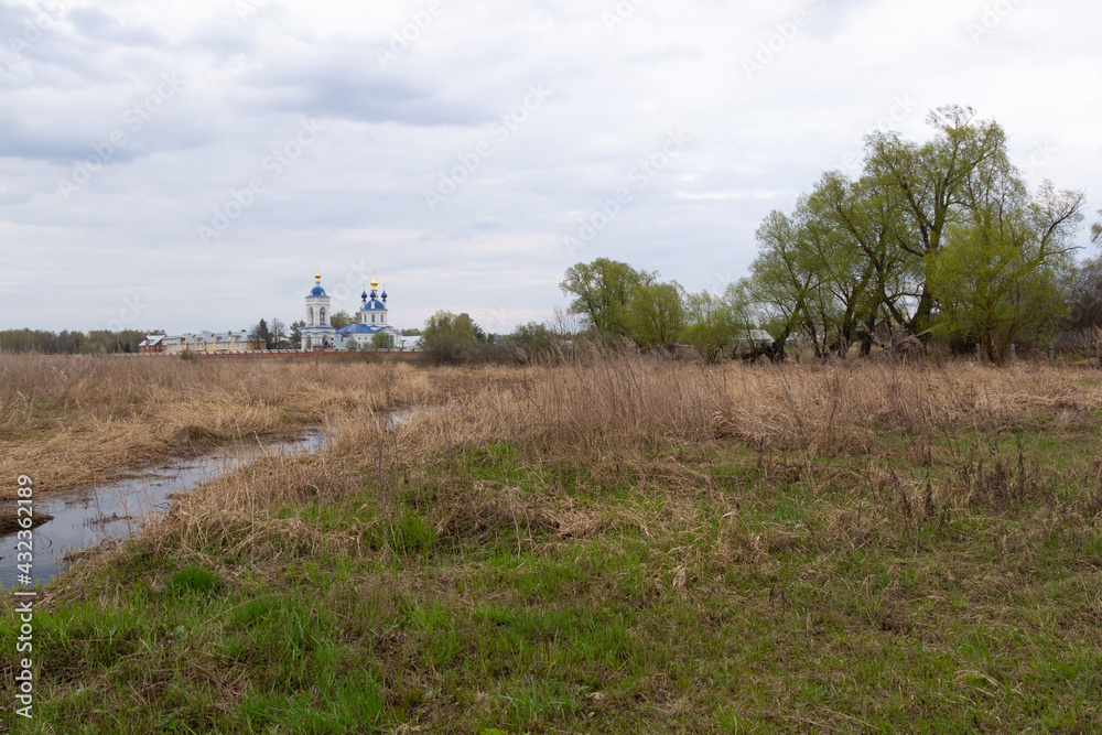 White and blue monastery in russian village Dunilovo, river, green field, trees in spring cloudy day