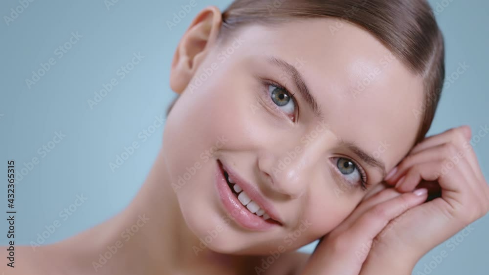 Headshot of pretty young woman with hands near her face looking to camera and leaning head . Portrait of female model with naked shoulders and light nude make up posing and smiling.
