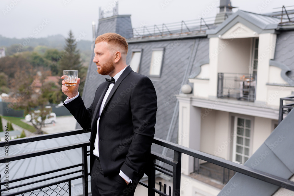 Side view of red-haired groom drinks whiskey on the balcony of the wedding morning. Concept of groom morning Side view of red-haired groom drinks whiskey on the balcony of the wedding morning. Concept of groom morning