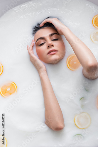 top view of woman with closed eyes relaxing in milky bath with sliced citrus fruits.