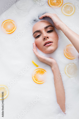 young woman with hands near face relaxing in milk bath with sliced citrus fruits.