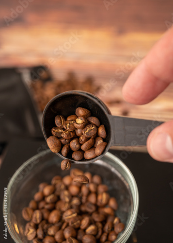 Weight measure, roasted coffee beans. Male hand pouring coffee beans from scoop into measuring glass cup, black bag of coffee in shallow background with coffee beans showing