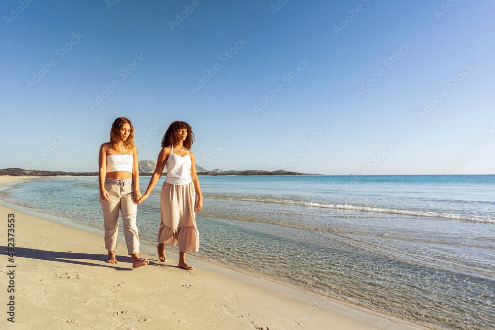 Multiracial couple of beautiful young gay women holding hands walking at sunrise or sunset on a tropical colored beach with flat ocean sea water. Concept: in love and nature there are no differences