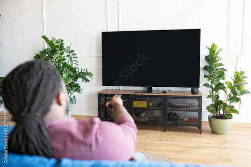 Back view African-American guy with dreadlocks holds remote controller and switching tv channels, watching TV sitting on the couch at home, the lcd TV with empty black screen, copy space, mockup