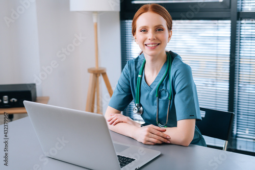 Portrait of attractive smiling young female doctor in blue green medical uniform sitting at desk with laptop on background of window in hospital office of medic clinic, looking at camera.