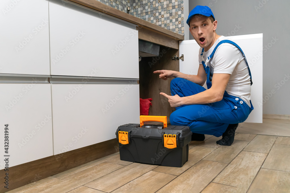 Handyman in overalls looks in surprise at the pipes under the sink in the kitchen, next to a box of tools Handyman in overalls looks in surprise at the pipes under the sink in the kitchen, next to a box of tools