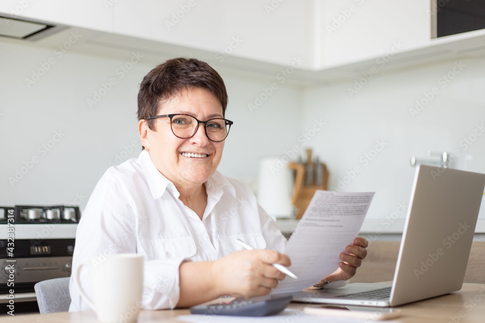 Smiling aged woman holding documents, checking information at laptop online, concerned senior female managing bank insurance or loan papers, busy working at computer. 