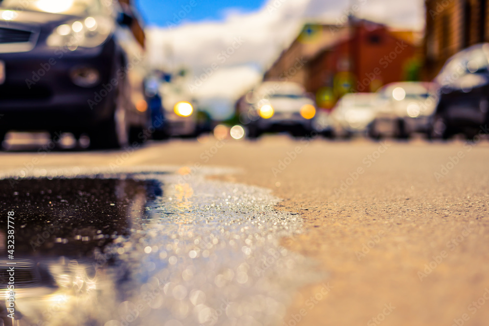 Sunny day after rain in the city, the car rides along the road. Close up view from the level of the puddle on the pavement Sunny day after rain in the city, the car rides along the road. Close up view from the level of the puddle on the pavement