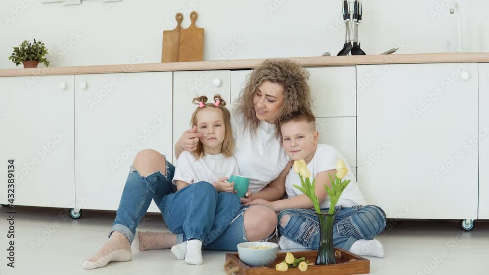 A tender family moment at home. A boy, a girl and a mother sit on the floor in a stylish white modern kitchen in Scandinavian design and have fun. Caucasian family in white T-shirts and jeans A tender family moment at home. A boy, a girl and a mother sit on the floor in a stylish white modern kitchen in Scandinavian design and have fun. Caucasian family in white T-shirts and jeans