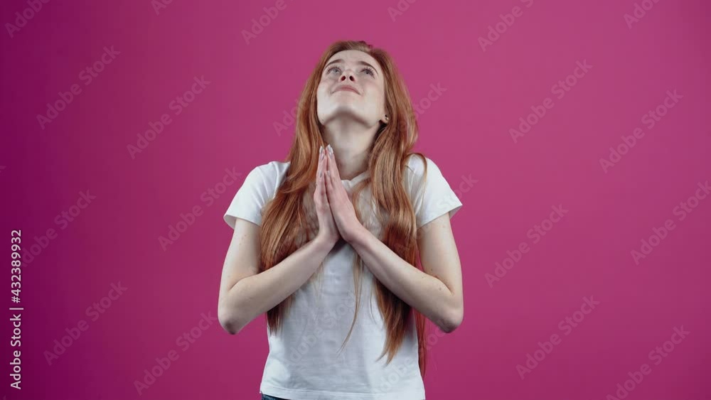PLEASE. The young redhead hopefully brings her hands together to her chest. Freckled teenage girl in a white T-shirt, isolated on a pink background. The concept of people's lifestyle.