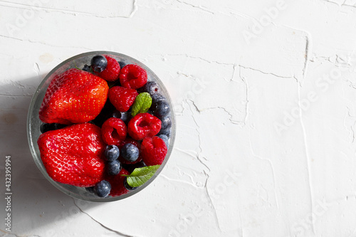 Fototapeta Naklejka Na Ścianę i Meble -  Appetizing variety of forest fruits (strawberries, large strawberries, raspberries, blueberries, red berries) in a glass bowl and mint leaves. On white textured background. Top view with copy space.