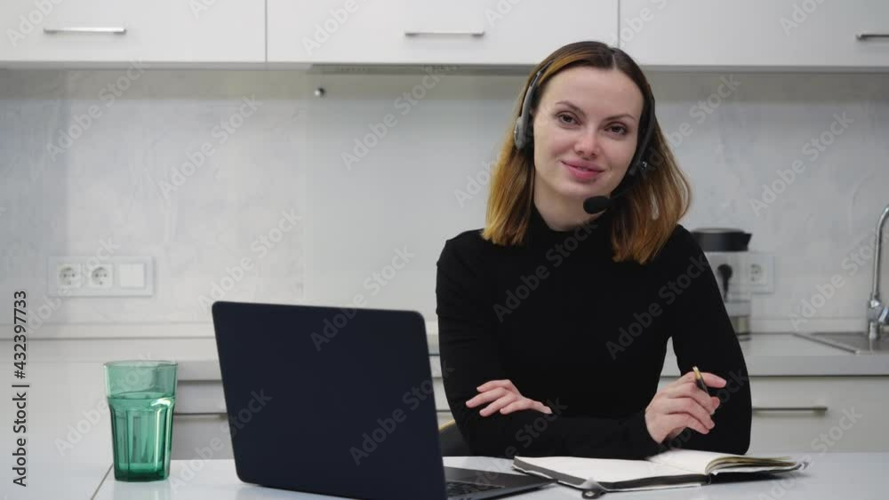 Attractive young girl in headset sits behind laptop, smiling straight at the camera. High quality 4k footage Attractive young girl in headset sits behind laptop, smiling straight at the camera. High quality 4k footage