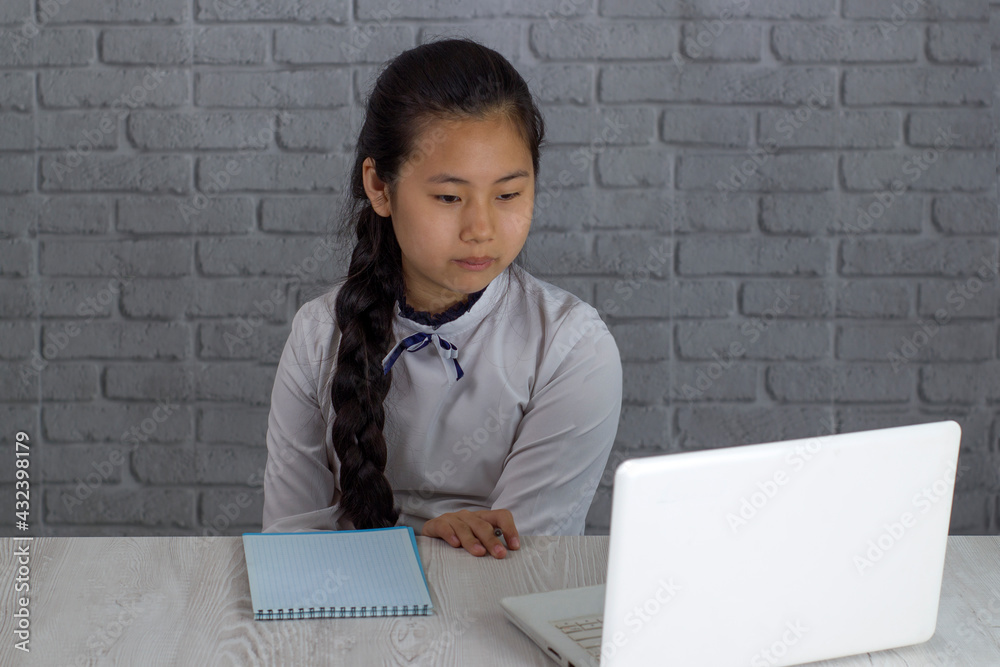 Little Asian girl studying at home in online class, learning online video call. She listens intently to an online school teacher with a laptop and takes notes at home. New normal