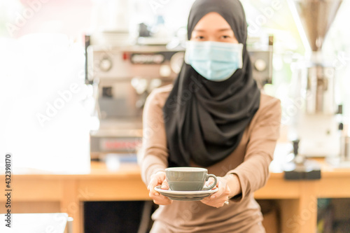 Beautiful Muslim woman wear mask for protect virus , cafe owner or clerk preparing to serve coffee to customers in the shop with a backdrop of equipment and coffee machines , focus on cup