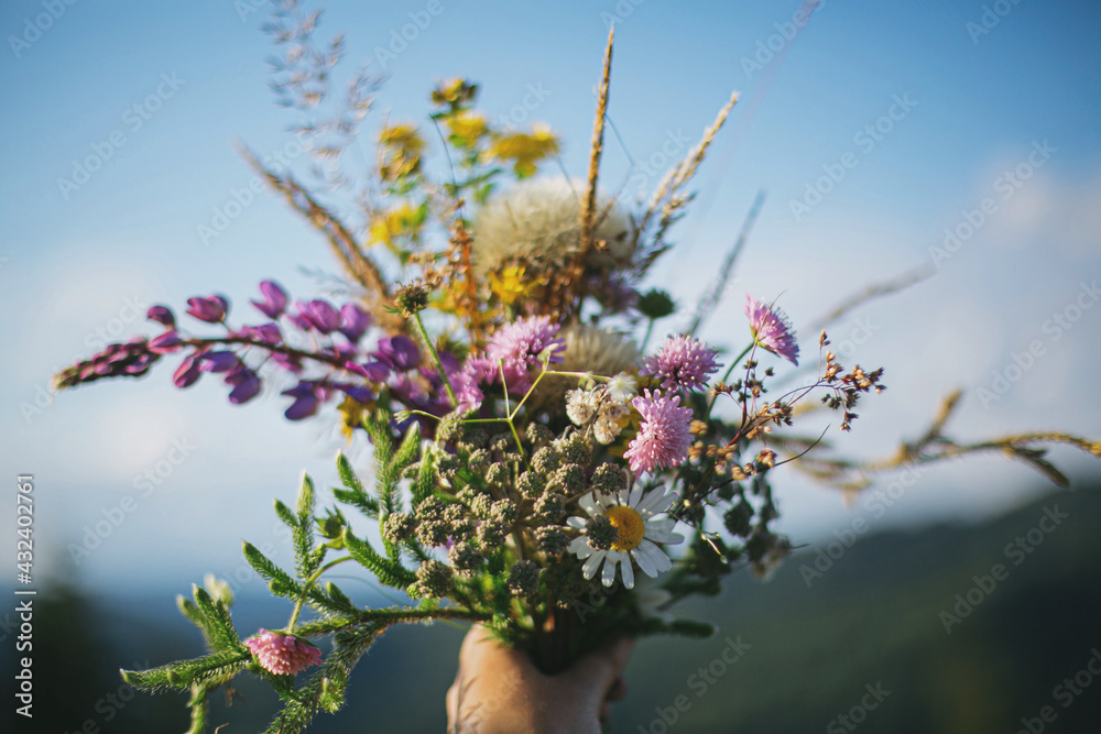 Beautiful colorful bouquet of wildflowers in hand on background of mountain hills and sunny sky - obrazy, fototapety, plakaty 