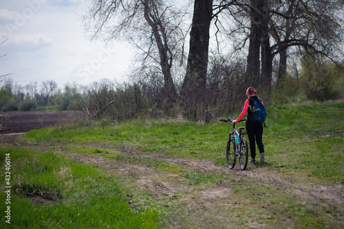 a girl with a bicycle walks a field trail. Woman riding a mountain bike in the forest. Hiker woman with backpack. one young woman riding a bike in nature. rolling the bike in his hands. back view.