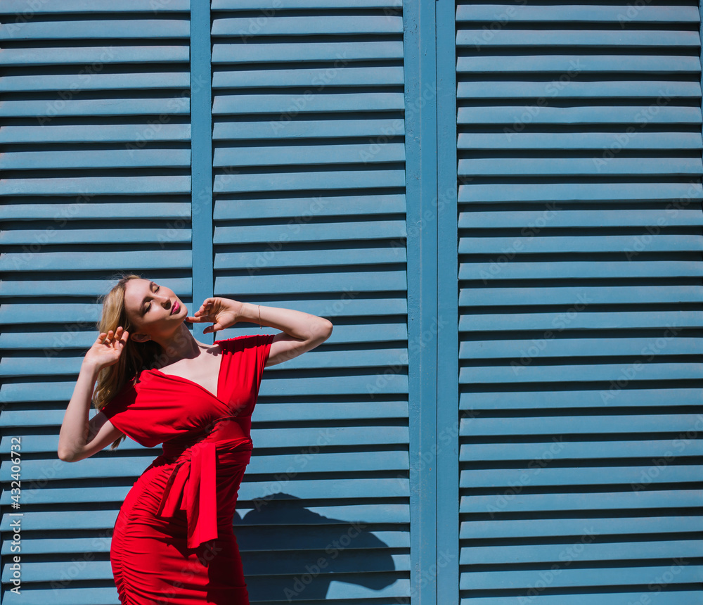 A young woman stands near a wall with blue shutters. Bright sunny day, hard light. A young woman stands near a wall with blue shutters. Bright sunny day, hard light.