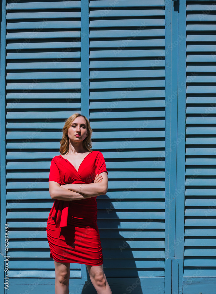 A young woman stands near a wall with blue shutters. Bright sunny day, hard light. A young woman stands near a wall with blue shutters. Bright sunny day, hard light.