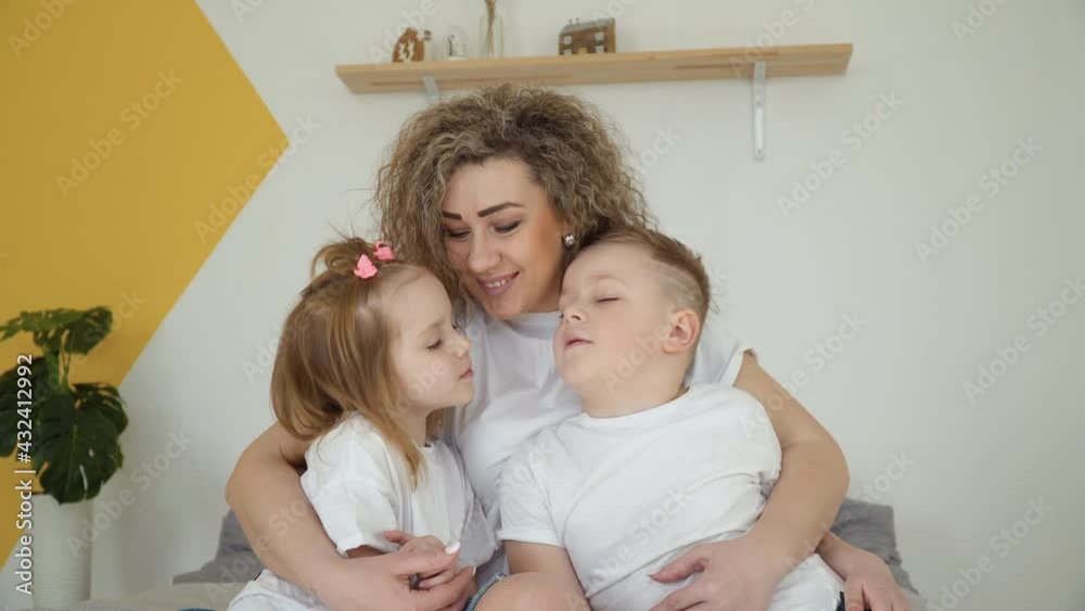 Daughter and son sit in mom's arms and kiss mom's cheeks. Family in a cozy room. Beautiful family portrait. White and yellow colors. Minimalism Daughter and son sit in mom's arms and kiss mom's cheeks. Family in a cozy room. Beautiful family portrait. White and yellow colors. Minimalism