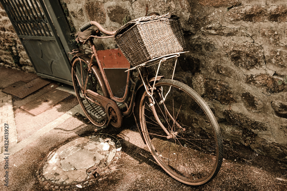 Rusty vintage bicycle with black board for entering a text (advertisement, menu etc) and wicker basket leaning on a stone wall. Brittany, France. Sepia toned photo.