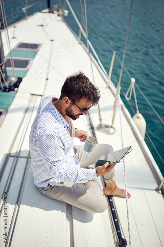 A young man is enjoying a coffee while sitting on a yacht at sea and using a tablet. Summer, sea, vacation
