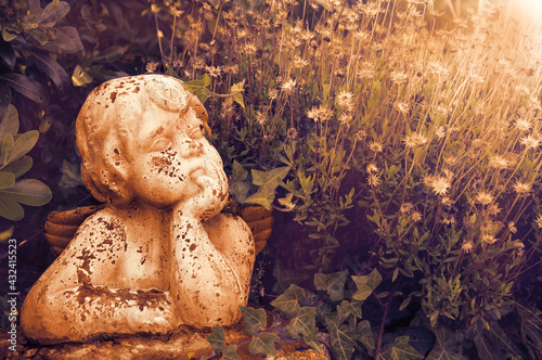 Canvas Print Weathered statue of an infant angel in overgrown garden in sunset golden light