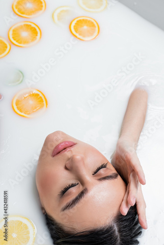 young woman with closed eyes touching face while relaxing in milk bath with citruses.