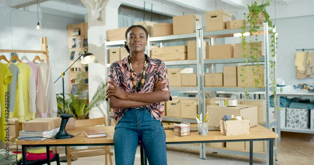 Cheerful positive African American young beautiful businesswoman with dreamy face standing in office in own small clothing store and looking at camera smiling in warehouse, stylist, business concept Cheerful positive African American young beautiful businesswoman with dreamy face standing in office in own small clothing store and looking at camera smiling in warehouse, stylist, business concept
