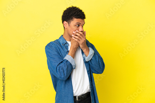 Young African American man isolated on yellow background covering mouth and looking to the side