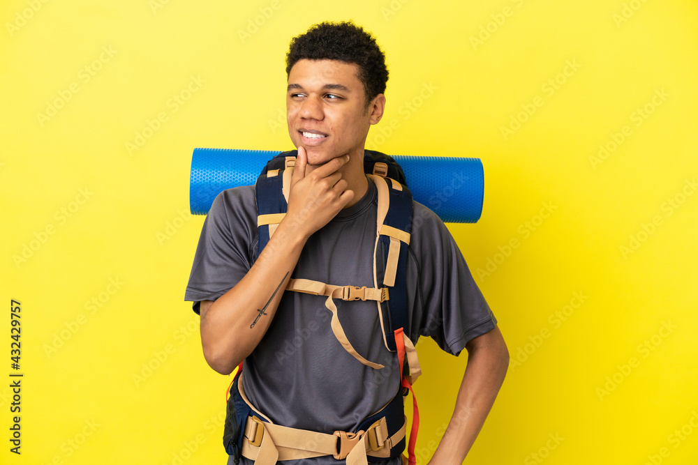 Young mountaineer African American man with a big backpack isolated on yellow background looking to the side and smiling
