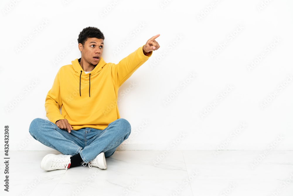 Young African American man sitting on the floor isolated on white background pointing away
