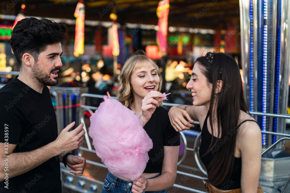 Dos chicas y un chico amigos sonriendo y compartiendo un algodón de azúcar rosa en zona de atracciones de una feria
