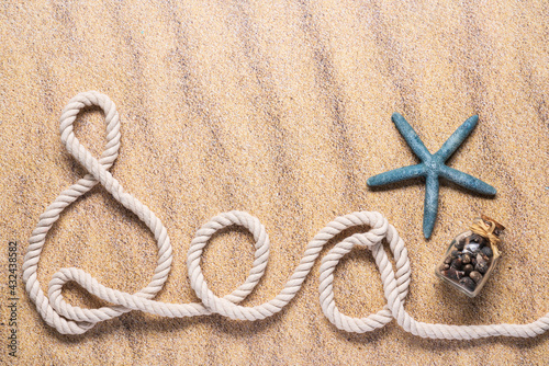 Sea spelled out with nautical rope with a blue starfish and small glass bottle of seashells on a beach sand background