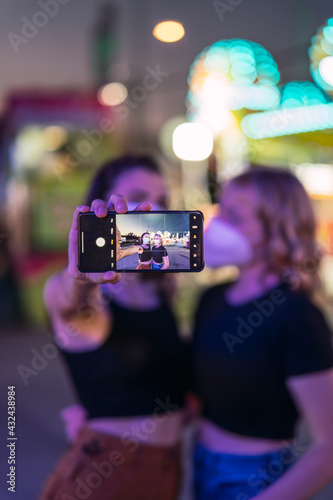 Dos chicas rubia y morena tomando selfies en zona de atracciones de una feria