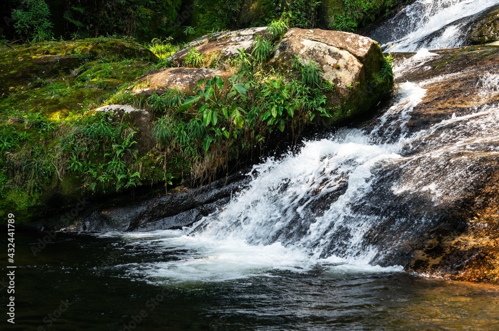 Naklejka premium Water current flowing around rock formations covered in moss of Ipiranguinha waterfall forming a golden natural pool right below in Serra do Mar (Sea Ridge) forest in Cunha, Sao Paulo - Brazil.