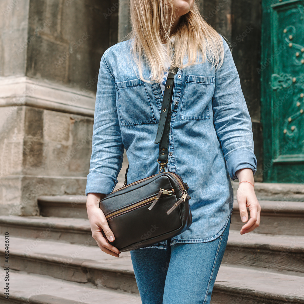Leather sling bag for women. Belt Bag Minimalist. Blond woman in denim  shirt and, blue jeans and a dark brown leather bag over the shoulder.  Close-up. Stock Photo | Adobe Stock