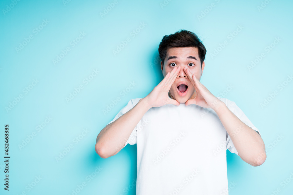 Young asian guy puts his hands around his mouth and opens it in surprise while standing against a bright background Young asian guy puts his hands around his mouth and opens it in surprise while standing against a bright background